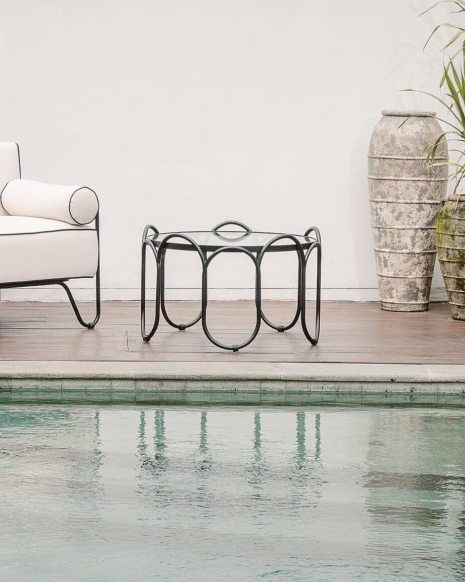 Outdoor seating area by a pool with white chairs and a small table, surrounded by greenery.