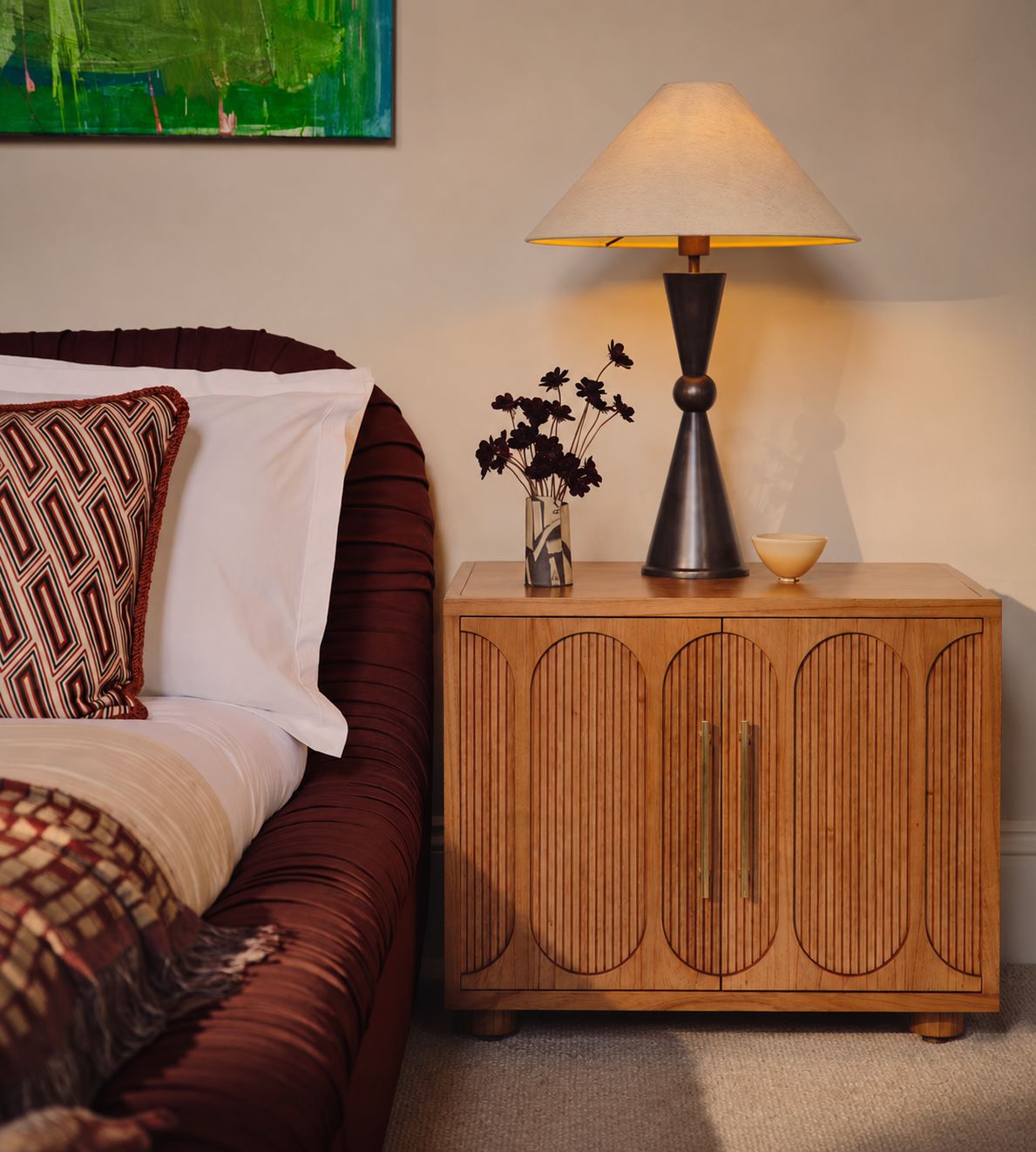 Wooden nightstand with lamp and decorative items next to a bed with patterned pillows.